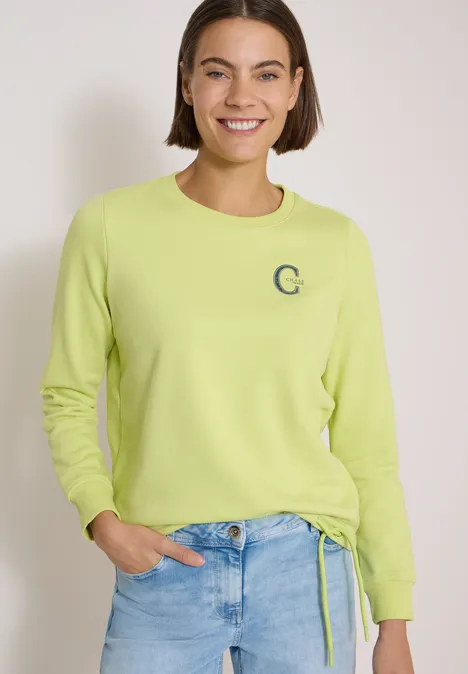 Fröhliche Frau mit Kurzhaarschnitt trägt gelb-grünen Pullover und helle Jeans.