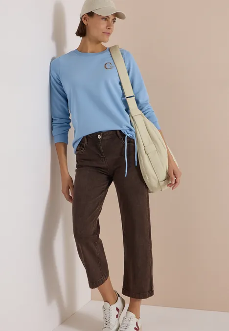 Frau in hellblauem Pullover, braunen Jeans und weißen Sneakers, mit weißer Cap und Stofftasche vor beiger Wand.