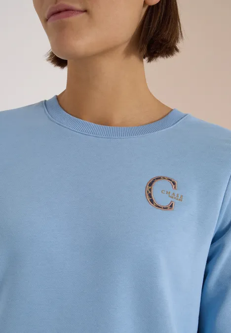 Porträt einer Person mit blauem Sweatshirt, auf der Brust ein goldfarbenes, verziertes 'C'-Logo.