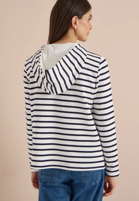Eine Frau von hinten, trägt einen weißen Kapuzenpullover mit dunkelblauen Streifen und Jeans.