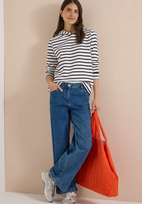 Frau in gestreiftem Langarmshirt, weitem Jeans, weißen Sneakers und orangefarbener Tasche vor neutralem Hintergrund.