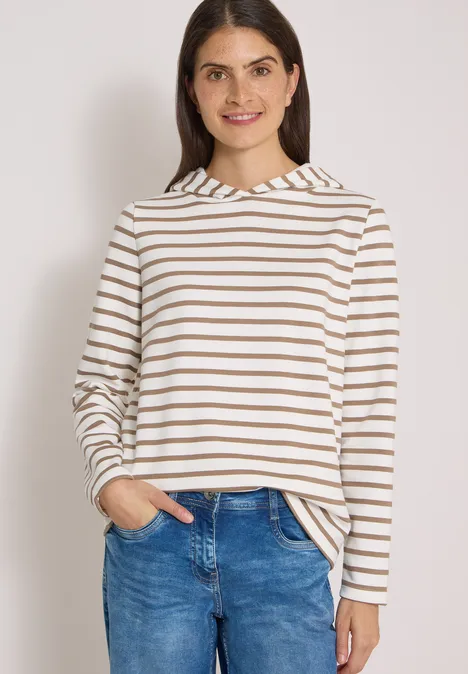 Frau in gestreiftem Langarmshirt (Weiß-Braun) und blauer Jeans, lächelt mit Händen in den Taschen.