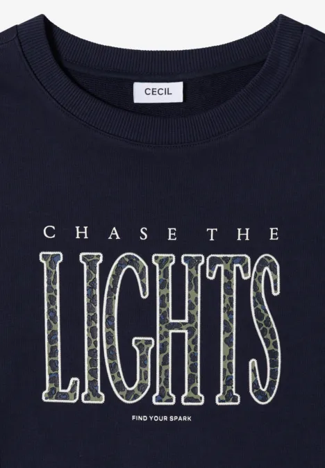 Schwarzes T-Shirt mit großem Slogan „CHASE THE LIGHTS FIND YOUR SPARK“ in weißen, dekorativen Buchstaben.
