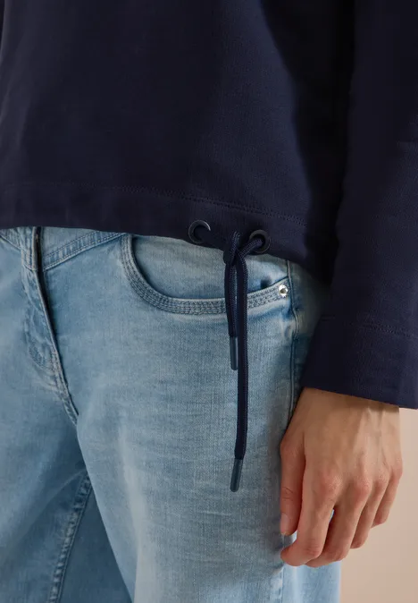 Dunkelblauer Pullover mit Kordelzug am Saum, dazu helle Jeans, Hand am Oberschenkel.