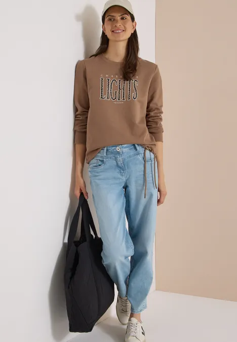 Frau in Jeans, braunem Pullover mit Aufdruck 'LIGHTS', Cap und schwarzer Tasche lehnt an weißer Wand.