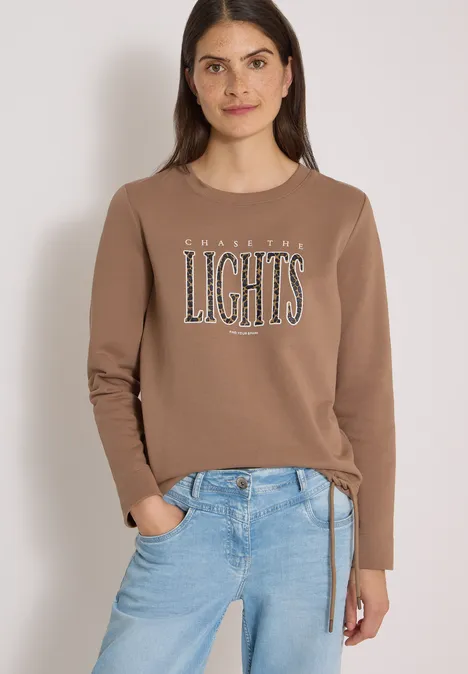 Eine Frau in braunem Sweatshirt mit Schriftzug ‘AMAZING LIGHTS’ und blauen Jeans vor hellem Hintergrund.