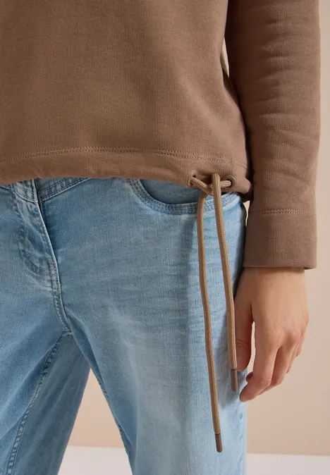 Eine Person trägt eine braune Pulloverjacke mit Tunnelzug und hellblaue Jeans; die linke Hand ruht locker an der Seite.
