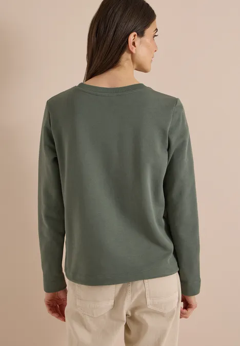 Frau von hinten in olivgrünem Langarmshirt und beigen Hosen vor hellem Hintergrund.