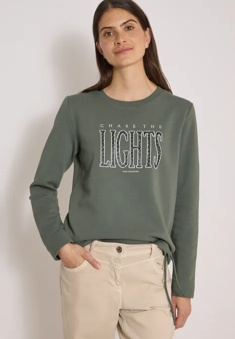 Frau in olivgrünem Sweatshirt mit Aufdruck 'LIGHTS' und hellen, hochgekrempelten Hosen, lässig stehend.