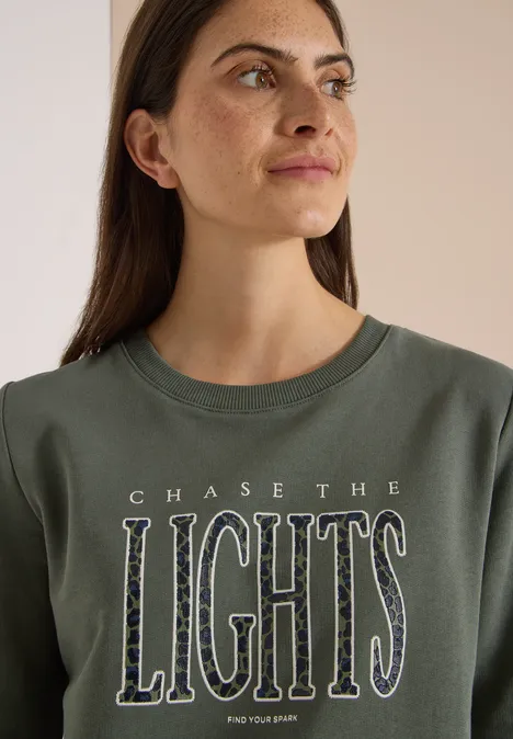 Frau mit langen dunklen Haaren trägt grünes Shirt mit Aufschrift 'CHASE THE LIGHTS'. Sie blickt seitlich.