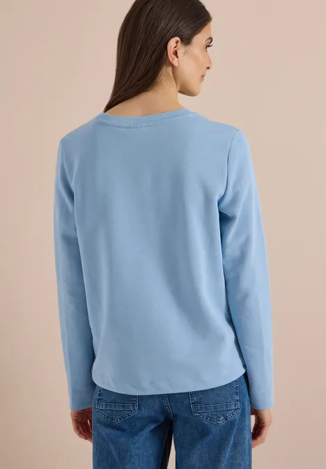 Frau von hinten in hellblauem Langarmshirt und Jeans vor neutralem Hintergrund.