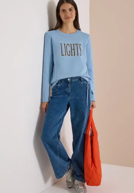 Frau in hellblauem Shirt mit Aufdruck 'LIGHTS', weiter Jeans, grauen Sneakern und oranger Tasche.
