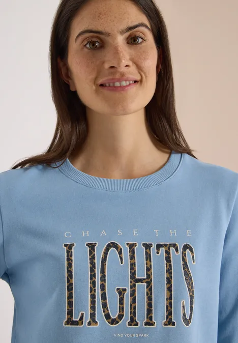 Frau mit langen braunen Haaren und Sommersprossen trägt hellblaues Shirt mit Aufschrift 'CHASE THE LIGHTS'.