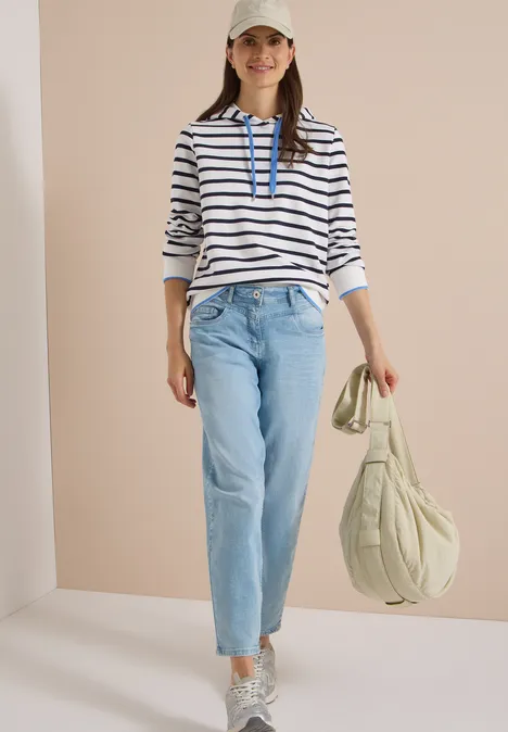 Frau in gestreiftem Kapuzenpullover, hellen Jeans und Cap hält eine leichte Tasche und trägt Sneaker.