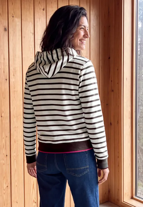 Sweatshirt mit Kapuze und Streifen truffle brown Eine Frau in gestreiftem Kapuzenpullover und Jeans steht vor einem Holzfenster und lächelt nach draußen.