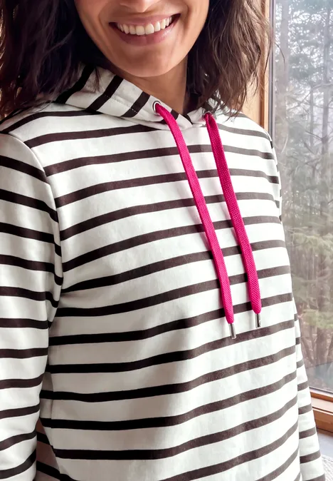 Sweatshirt mit Kapuze und Streifen truffle brown Nahaufnahme einer Frau mit gestreiftem Kapuzenpullover und pinken Kordeln, die lächelnd posiert.