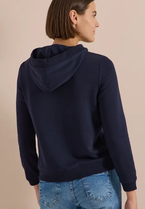 Rückansicht einer Frau mit kurzem Haar, sie trägt einen dunkelblauen Kapuzenpullover.