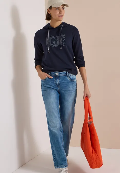 Frau in Navy-Hoodie, weiten Jeans und Basecap, trägt eine orangefarbene Tasche.