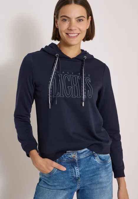 Frau in dunkelblauem Hoodie mit 'NIGHTS'-Aufdruck und Jeans lächelt entspannt in die Kamera.