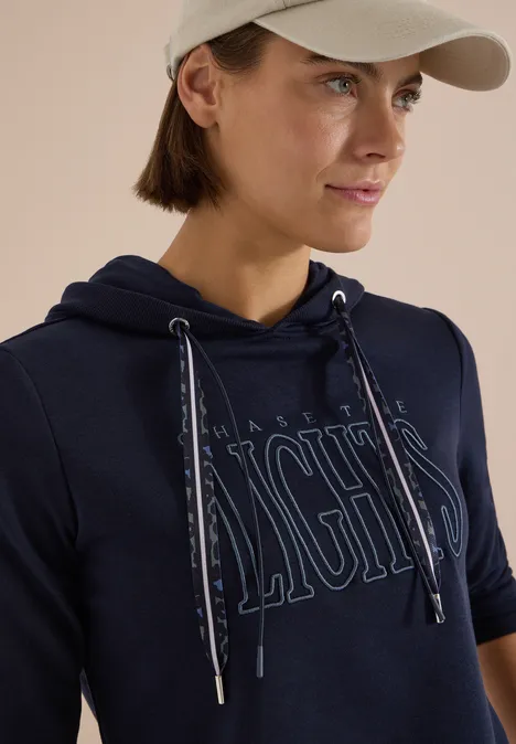 Frau in dunkelblauem Hoodie mit modischem Schriftzug und heller Cap, Nahaufnahme.