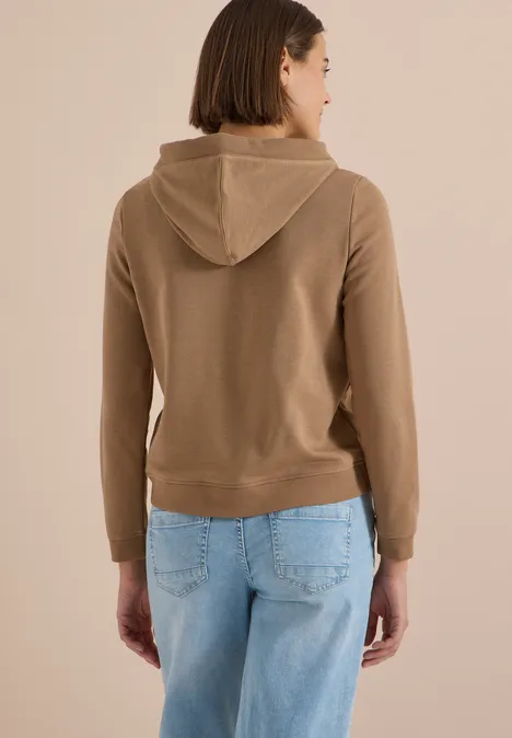 Frau von hinten, trägt beigefarbene Kapuzenjacke und hellblaue Jeans.