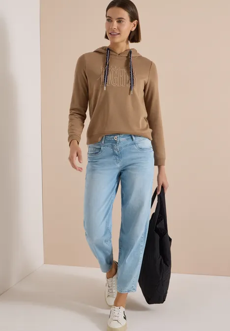 Frau in brauner Kapuzenjacke, hellen Jeans und weißen Sneakern trägt eine große schwarze Tasche.