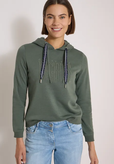 Junge Frau in olivgrünem Hoodie mit Kordelzug und hellen Jeans, lächelt vor neutralem Hintergrund.
