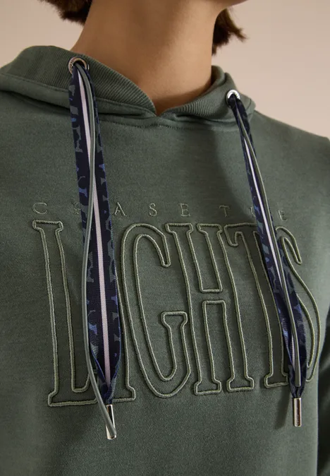 Grüner Hoodie mit geprägtem ‚CHASE LIGHTS‘-Schriftzug und gemusterten Kordeln, nur Oberkörper sichtbar.