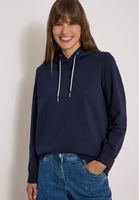Een vrouw met bruin haar draagt een donkerblauwe hoodie en jeans, glimlachend voor een lichte achtergrond.
