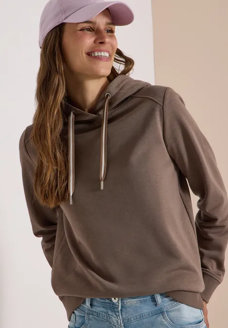Jonge vrouw lachtend in een hoodie, spijkerbroek en pet, zijwaarts kijkend.