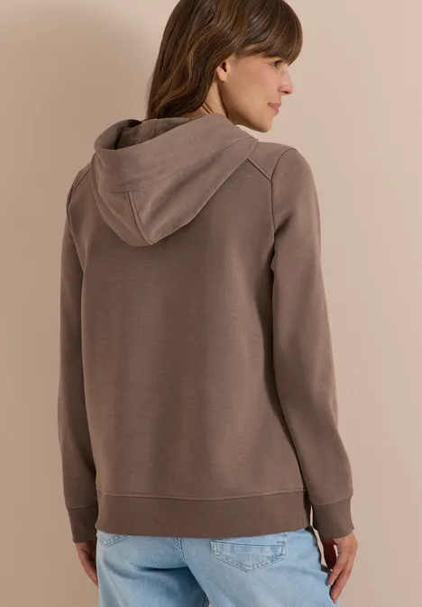 Vrouw van achteren in een bruine hoodie en blauwe jeans, licht zijwaarts gedraaid.