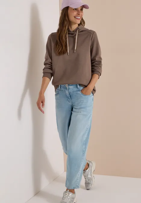 Jonge vrouw draagt bruine hoodie, blauwe jeans en witte sneakers, leunt tegen de muur.