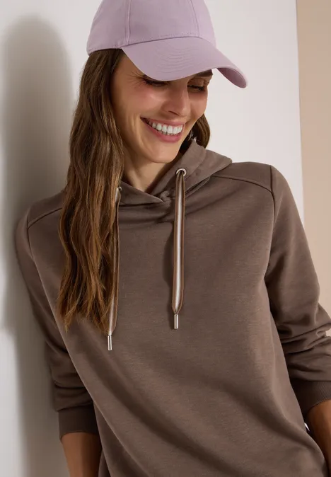 Vrouw lacht in een bruine hoodie en paarse pet.