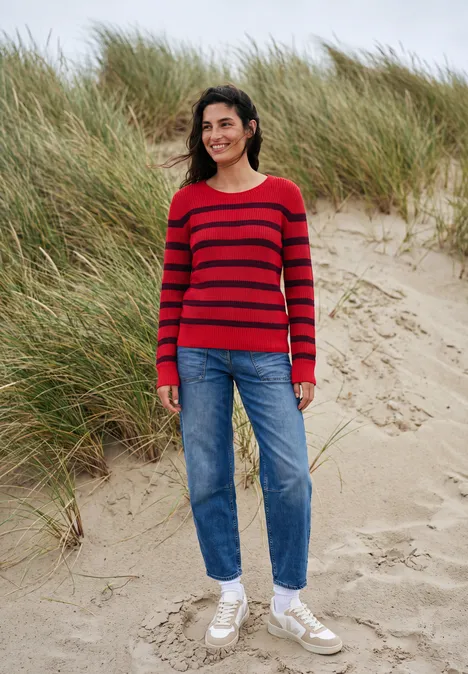 Vrouw in rood gestreepte trui, jeans en witte sneakers staand en lachend op een zandduin met gras.