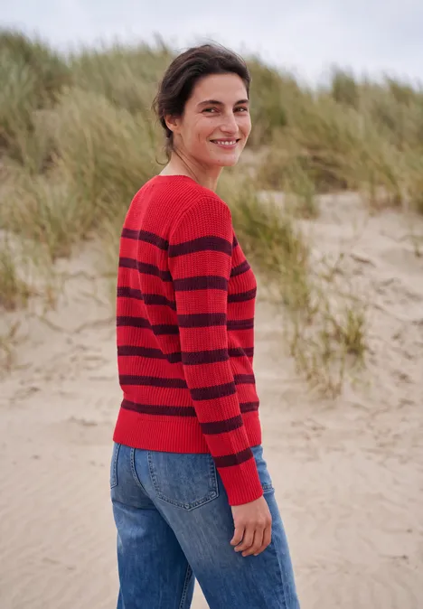 Een glimlachende vrouw in een rood-zwart gestreepte trui staat op het strand voor duinen.