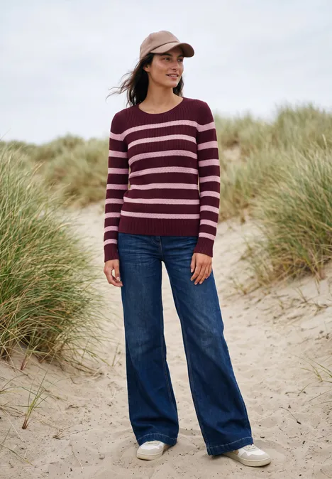 Vrouw met gestreepte trui, wijdflared jeans en pet, glimlachend tussen de duinen aan zee.