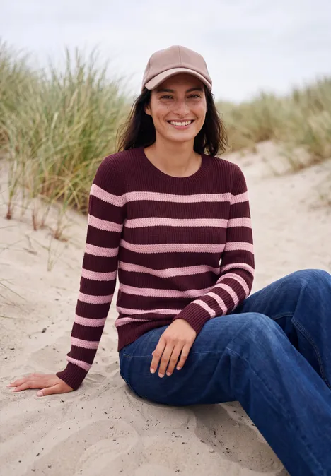 Een lachende vrouw zit op het strand in jeans, gestreepte trui en pet, met duinen op de achtergrond.