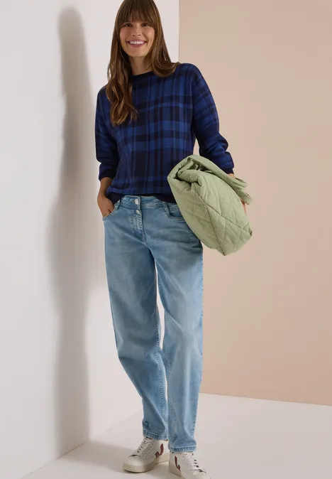 Dolman Pullover mit Karomuster urban dark blue Frau in blauem Karo-Pullover, heller Jeans und Sneakern, die eine grüne Steppjacke hält und lächelt.