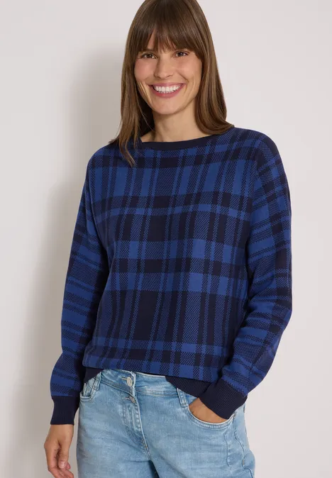 Dolman Pullover mit Karomuster urban dark blue Frau lächelt, trägt blau karierten Pullover und Jeans vor neutralem Hintergrund.