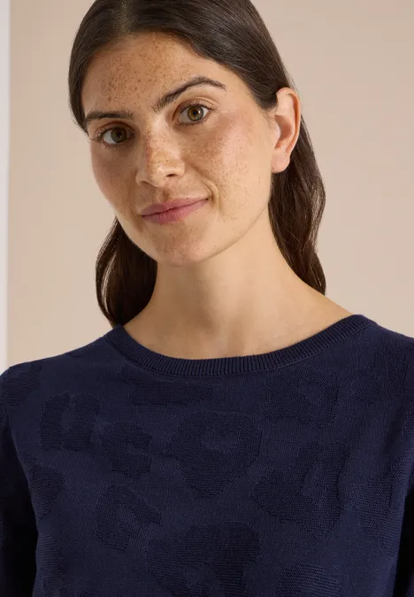 Pull à col bateau et texturé urban dark blue Porträt einer Frau mit dunklen Haaren und Sommersprossen in marineblauem Pullover vor beigem Hintergrund.