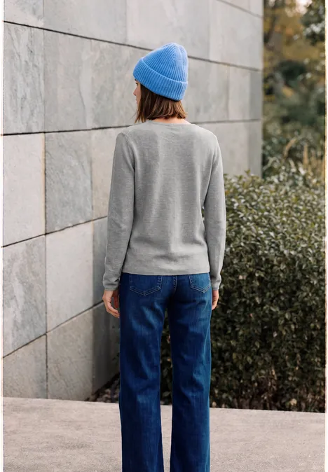Person in grauem Pullover, blauer Mütze und Jeans steht mit Rücken zur Kamera vor einer Mauer.