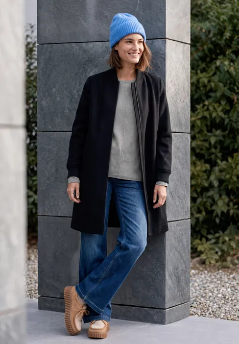 Eine Frau in schwarzem Mantel, Jeans, grauem Pullover und blauer Mütze lehnt lächelnd an einer Säule im Freien.