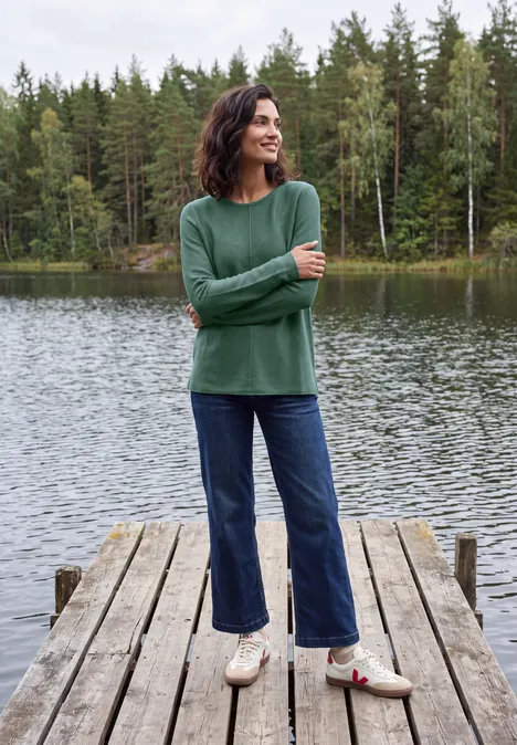 Frau im grünen Pullover und Jeans steht auf einem Steg an einem ruhigen See mit Wald im Hintergrund.