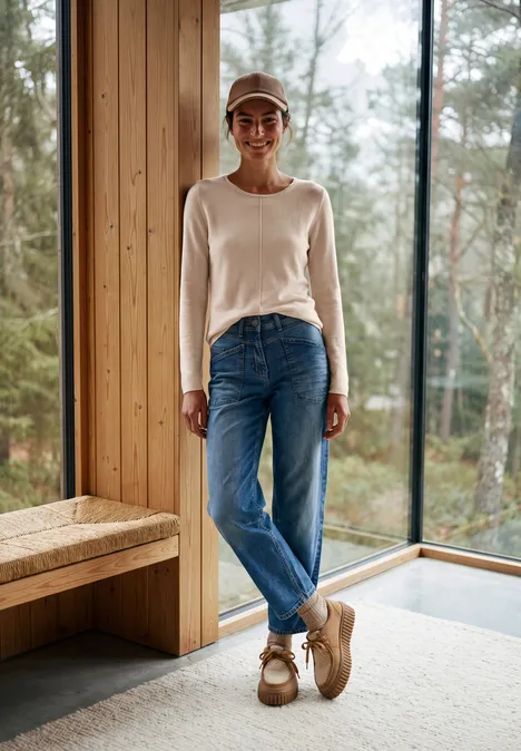 Vrouw in jeans en licht trui lacht staand in een houten huis voor grote ramen, met uitzicht op het bos.