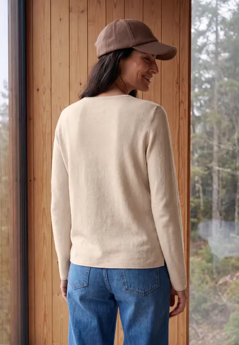Een vrouw in een beige trui, blauwe jeans en bruine pet staat in een houten kamer voor bosraam.