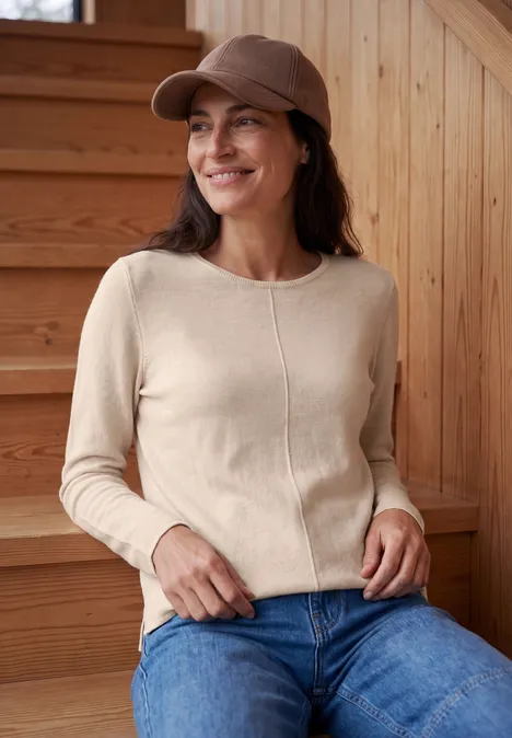 Vrouw met beige trui, bruine pet en jeans zit glimlachend op een houten trap.