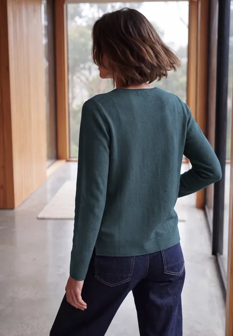 Vrouw van achteren in een groen trui en blauwe jeans bij grote ramen.