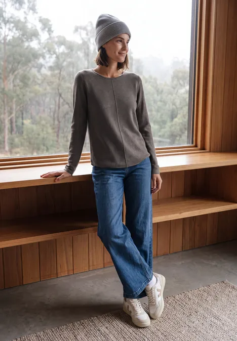 Frau in grauem Pullover und Jeans lehnt entspannt am Fenster aus Holz, trägt Mütze und Sneakers.