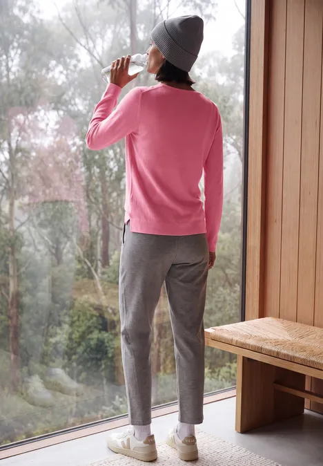 Eine Person in rosa Pullover, grauer Mütze und Hose steht an einem Fenster und trinkt aus einer Tasse.
