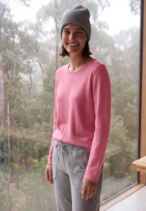 Eine lächelnde Frau im rosa Pullover und grauen Hosen mit Mütze am Fenster, Wald im Hintergrund.